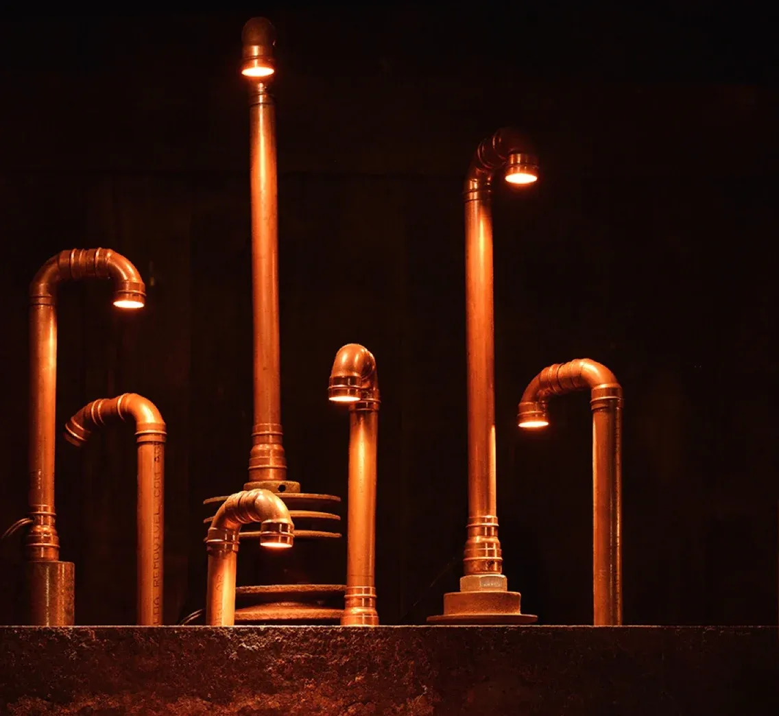 Seis luminárias de tubo de cobre acesas sobre bancada de aço enferrujado